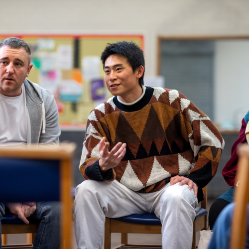 Man in brown jumper sitting on a chair speaking to people in a circle